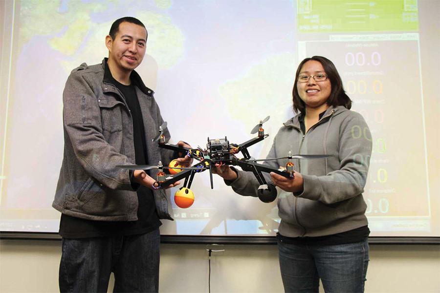 Two individuals stand in front of a screen while holding a drone